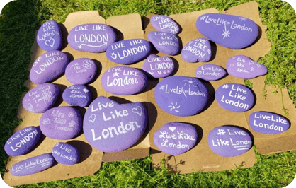 Dozens of hand-painted purple Live Like London rocks arranged on a board on grass.