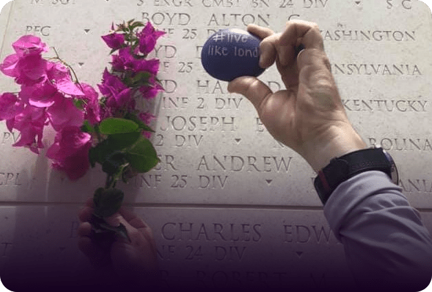 A purple Live Like London rock placed at a military memorial wall beside bougainvillea flowers.